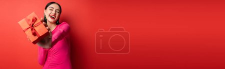Téléchargez les photos : Charmante femme avec des cheveux bruns et des boucles d'oreilles à la mode souriant tout en se tenant dans la robe de fête magenta et tenant cadeau enveloppé pour les vacances sur fond rouge, bannière - en image libre de droit