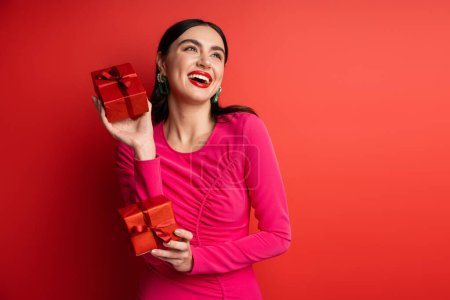 overjoyed woman with brunette hair and trendy earrings smiling while standing in magenta party dress and holding wrapped gift boxes for holiday on red background 
