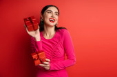 overjoyed woman with brunette hair and trendy earrings smiling while standing in magenta party dress and holding wrapped gift boxes for holiday on red background  Poster #654500880