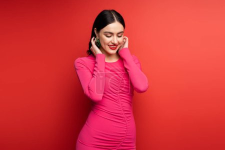 glamorous woman with brunette hair and trendy earrings smiling while standing in magenta party dress while posing and looking down on red background