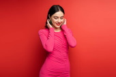 glamorous woman with brunette hair and trendy earrings smiling while standing in magenta party dress while posing and looking down on red background mug #654501038