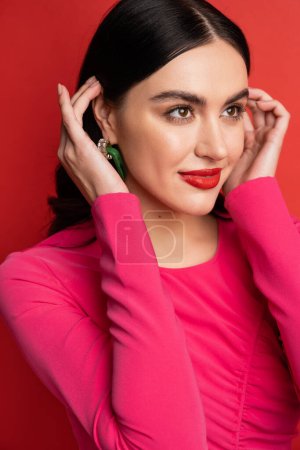 Photo for Portrait of alluring woman with brunette hair and trendy earrings smiling while standing in magenta party dress while posing and looking away on red background - Royalty Free Image