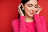 joyful and shy woman in trendy earrings adjusting her brunette hair and smiling while standing in magenta party dress while posing on red background Sweatshirt #654501062