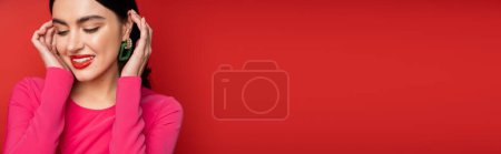 mujer glamorosa con cabello moreno y pendientes de moda sonriendo mientras está de pie en vestido de fiesta magenta y posando, mirando hacia otro lado sobre fondo rojo, pancarta 