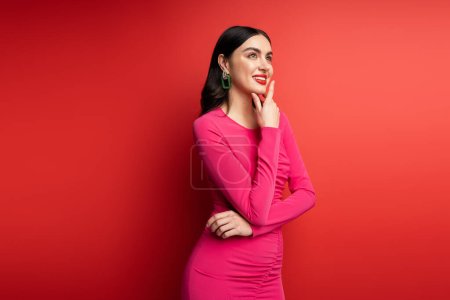 pretty woman with brunette hair and trendy earrings smiling while standing in magenta party dress while posing and looking way, thinking on red background
