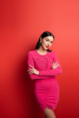 Photo for Gorgeous woman with brunette hair and trendy earrings standing with folded arms in magenta party dress while posing and looking at camera on red background - Royalty Free Image