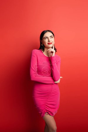 charming woman with brunette hair and trendy earrings smiling while thinking and holding hand near face and standing in magenta party dress on red background