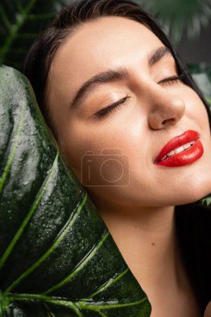 vista de cerca de la seductora joven con el pelo moreno y los labios rojos sonriendo mientras posa con los ojos cerrados alrededor de las hojas de palmeras tropicales, húmedas y verdes con gotas de lluvia 