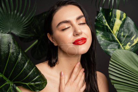 gorgeous young woman with brunette hair and red lips smiling while posing with closed eyes around tropical, wet and green palm leaves with raindrops on them  