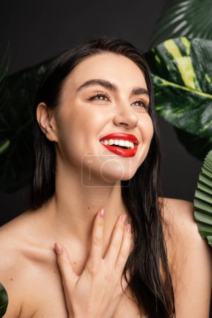glückliche junge Frau mit brünetten Haaren und roten Lippen lächelt, während sie mit der Hand auf der Brust um nasse und grüne Palmenblätter mit Regentropfen auf ihnen posiert und wegschaut