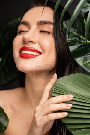 positive young woman with brunette hair and red lips smiling while posing with closed eyes around tropical, wet and green palm leaves with raindrops on them 