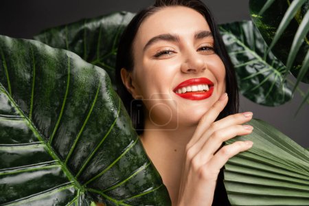 jeune femme positive avec les cheveux bruns et les lèvres rouges souriant tout en posant autour de feuilles de palmier vert exotique avec des gouttes de pluie sur eux et en regardant la caméra 