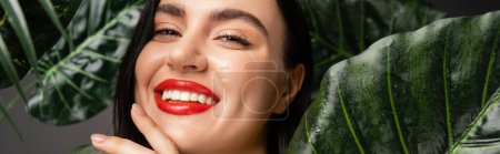 positive woman with brunette hair and red lips smiling while posing around exotic and green palm leaves with raindrops on them and looking at camera, banner 
