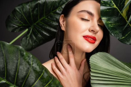 pleased young woman with brunette hair and red lips smiling while posing with closed eyes next to tropical green palm leaves with raindrops on them 