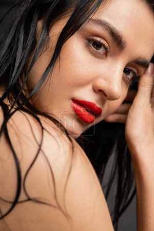 portrait of young and charming young woman with wet brunette hair and red lips posing with bare shoulder while looking at camera after shower isolated on grey background 