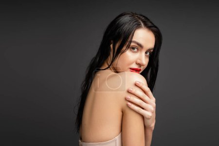 portrait of young and graceful woman with wet brunette hair and red lips touching bare shoulder while looking at camera after shower isolated on grey background 