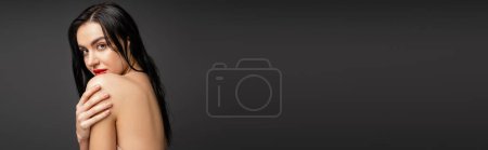 Téléchargez les photos : Portrait de jeune et jolie femme aux cheveux mouillés et bruns touchant épaule nue tout en regardant caméra après douche isolé sur fond gris, bannière - en image libre de droit
