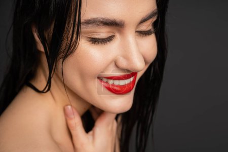 close up view of young and pretty woman with wet brunette hair, red lips posing with hand on neck and smiling after shower isolated on grey background 
