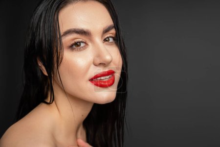 portrait of young and graceful woman with wet brunette hair and red lips posing while looking at camera after shower isolated on grey background 