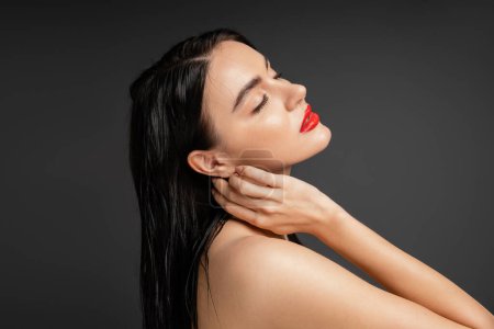 gorgeous and graceful woman with wet brunette hair and red lips touching neck while posing with bare shoulders after shower isolated on grey background 