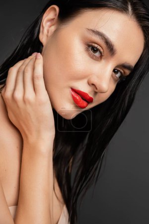portrait of young gorgeous and graceful woman with wet brunette hair and red lips touching neck while posing with bare shoulders and looking at camera isolated on grey background 