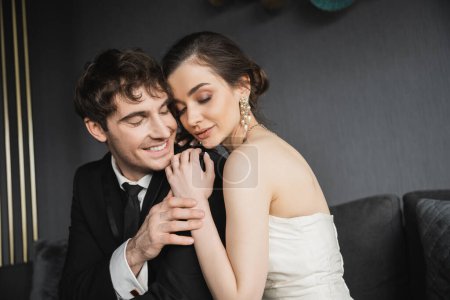 Porträt einer jungen brünetten Braut in luxuriösen Ohrringen mit Perlen und weißem Hochzeitskleid, die mit geschlossenen Augen sitzt und den glücklichen Bräutigam im schwarzen Anzug im Hotelzimmer umarmt 