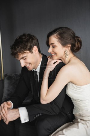 portrait of charming young bride in white wedding dress and handsome groom in black suit smiling together while sitting in hotel room, happy newlyweds