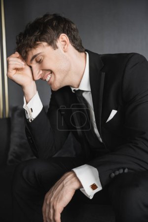 retrato de novio alegre y joven en traje negro con camisa blanca y corbata tocando la cara con la mano mientras sonríe y se sienta en un cómodo sofá en la habitación de hotel moderna el día de la boda