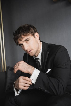 portrait of confident and young groom in black suit with white shirt and tie adjusting cufflinks and sitting on dark grey couch while looking at camera in modern hotel room on wedding day
