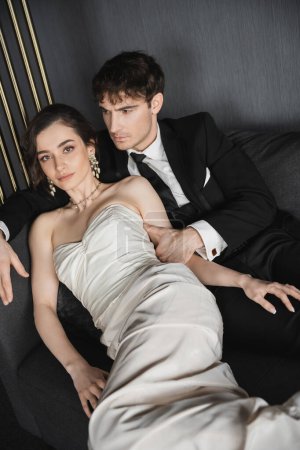 Charmante junge Braut in luxuriösen Ohrringen mit Perlen und weißem Hochzeitskleid lehnt an Bräutigam im schwarzen Anzug mit Krawatte, während sie zusammen auf dunkelgrauer Couch im Hotelzimmer sitzt