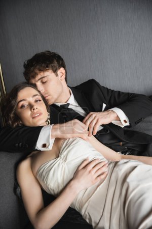 gut aussehender Bräutigam im schwarzen Anzug mit Krawatte umarmt und küssende Braut in Ohrringen mit Perlen und Halskette liegt im weißen Hochzeitskleid auf dunkelgrauer Couch im Hotelzimmer