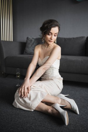 elegant bride with brunette hair sitting in white wedding dress, high heels and luxurious jewelry next to glass of champagne on floor near couch in hotel suite 