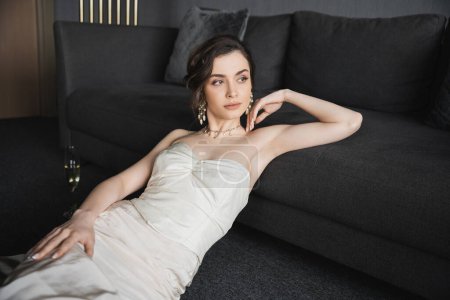 elegant bride with brunette hair posing in white wedding dress, luxurious jewelry, earrings with pearls and necklace next to glass of champagne on floor and leaning couch in hotel room
