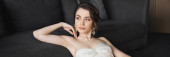 portrait of dreamy and gorgeous bride with brunette hair sitting in elegant and white wedding dress, luxurious jewelry, earrings and necklace and looking away in hotel room, banner  hoodie #654952670
