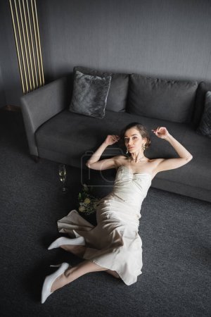 Blick auf die Braut mit brünetten Haaren im weißen Hochzeitskleid, High Heels und luxuriösem Schmuck neben einem Glas Champagner und einem Strauß auf dem Boden neben der Couch im Hotelzimmer