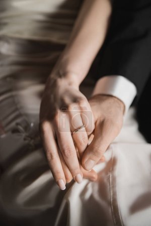 vista recortada de los recién casados, novia con elegante y lujoso anillo de bodas en el dedo y el novio en traje tomados de la mano el uno del otro después de la boda en la habitación de hotel 