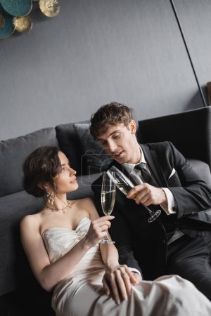 happy newlyweds, bride in white wedding dress and groom in black suit holding glasses of champagne while clinking and celebrating their marriage after wedding in hotel room  