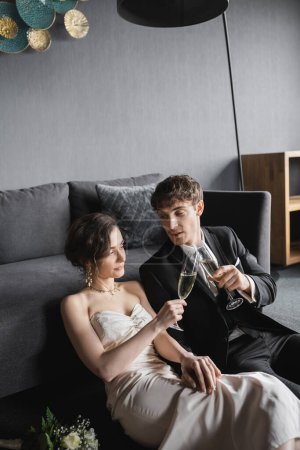 bride in elegant white wedding dress and groom in black suit clinking glasses of champagne while celebrating their marriage near bridal bouquet after wedding in hotel room  