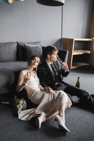 verträumte Braut im eleganten weißen Hochzeitskleid und Bräutigam im schwarzen Anzug mit Champagnergläsern, während sie ihre Hochzeit in der Nähe von Brautstrauß und Couch nach der Hochzeit im Hotelzimmer feiern  