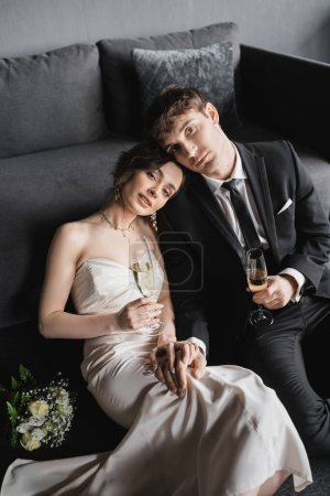 happy newlyweds, bride in white wedding dress and groom in black suit holding glasses of champagne while looking at camera after wedding ceremony and sitting near bridal bouquet in hotel room  