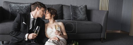 bride in white wedding dress and groom in black suit holding glasses of champagne while kissing and celebrating their marriage near bridal bouquet after wedding in hotel room, banner 
