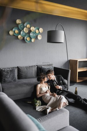 joyful bride in white wedding dress and groom in black suit holding glasses of champagne while celebrating their marriage near bridal bouquet after wedding in hotel room, happy newlyweds
