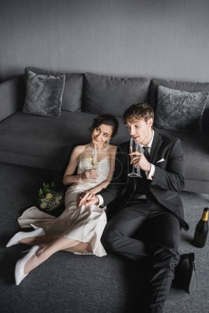 glückliche Braut im weißen Hochzeitskleid und Bräutigam im schwarzen Anzug mit Champagnergläsern, während sie ihre Hochzeit in der Nähe von Brautstrauß und Flasche nach der Hochzeit im Hotelzimmer mit Couch feiern 