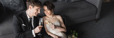 happy bride in white wedding dress and groom in black suit clinking glasses of champagne while celebrating their marriage near bridal bouquet after wedding in hotel room, banner 