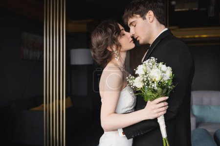 Photo for Side view of good looking groom in black formal wear kissing brunette bride in elegant wedding dress holding bridal bouquet with flowers while standing in hotel lobby - Royalty Free Image