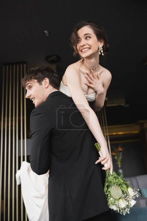 gut aussehender Bräutigam in schwarzer Festkleidung, der fröhliche Braut mit Make-up in weißem Hochzeitskleid mit Brautstrauß aus Blumen hebt, während er in der Hotellobby steht 