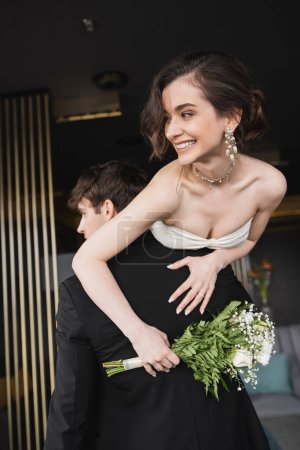 Photo for Groom in black formal wear lifting joyful bride in white wedding dress and luxurious jewelry holding bridal bouquet with flowers while standing in hotel lobby - Royalty Free Image