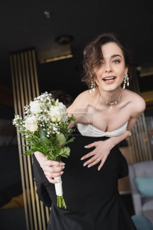 Bräutigam in schwarzer Festkleidung hebt hübsche Braut in weißem Hochzeitskleid und luxuriösem Schmuck und hält Brautstrauß mit Blumen in der Hotellobby 