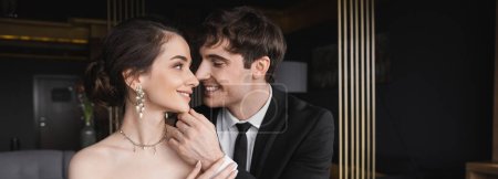 glücklicher Bräutigam im schwarzen Anzug mit Krawatte berührt Gesicht der charmanten Braut in Ohrring und Halskette, während sie einander in modernem Hotelzimmer anschauen, Banner 