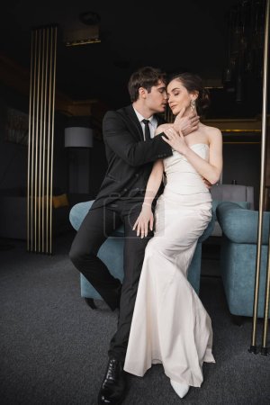 Zarter Bräutigam im schwarzen Anzug mit Krawatte berührt Gesicht der charmanten Braut im weißen Hochzeitskleid und Schmuck lehnt auf blauem Sofa im modernen Hotelzimmer